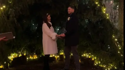 @reelallaboutit Sinead and Michael hold hands during their wedding ceremony which took place in a forest in front of an array of conifer branches and fairy lights