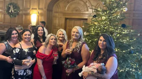 Victoria Walker Seven women and a baby pose in front of the Christmas tree set up in the wood-panelled venue where the Hollidays had their wedding