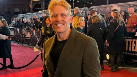 Adam Tucker Adam Tucker wearing a suit blazer and black t-shirt stood on the London red carpet premiere of Better Man. Fans and press are stood on the other side of a barrier behind him.