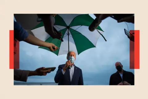 Getty Images Joe Biden under an umbrella with a mask on, as press ask questions 