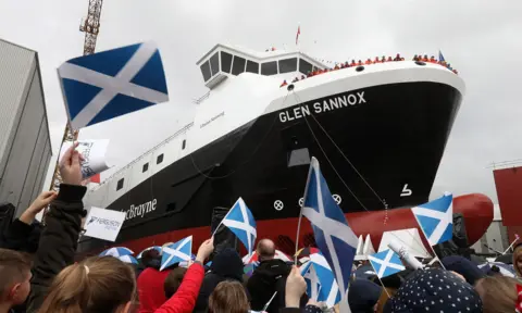 PA Media People wave saltires while watching a large red, black and white ship move down the slipway.