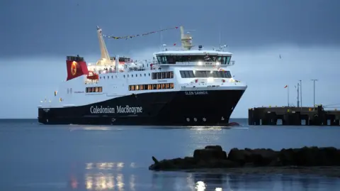 PA Media Glen Sannox beside a dock under a dark blue dawn sky. The boat is lit by yellow lights with a line of flags along the top.