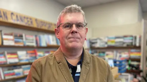 A grey haired bespectacled man in a brown jacket looks at the camera with second hand books in the background and a sign saying Arran Cancer Support