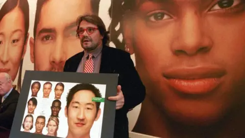 Getty Images Toscani wears a dark jacket and holds a display of models of different ethnicities he has photographed. He has long hair, and wears a red and white striped tie and red framed glasses. 
