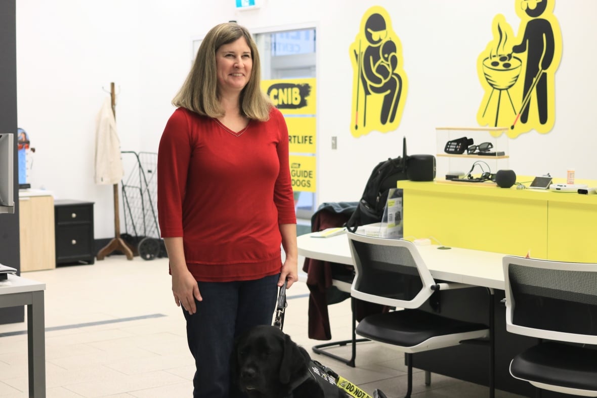 A woman with long hair stands in an office with her guide dog sitting at her feet. A sign in the background reads "CNIB."