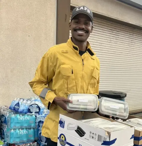 Fardad Khayami A man in a yellow jacket holds several trays of meals, as he wears an LA fire department hat, standing in front of a crate of bottled water