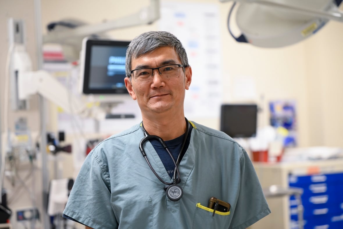 A man in scrubs and wearing a stethoscope smiles in a hospital room.