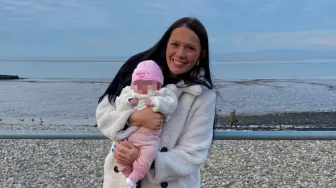 Lancashire Police Kiena Dawes, who has long, straight black hair and wears a furry white coat, smiles at the camera holding her baby daughter, who wears a pink hat with the words 'Mummy's Girl' stitched onto it. In the background is a pebbly beach.