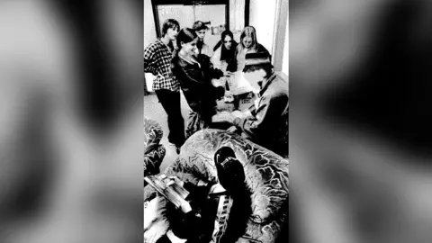 Peter de Boer A black and white photo of Banksy, wearing a bucket hat, surrounded by young people inside a youth centre.