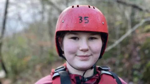 School pupil Elizabeth, 12, smiling at the camera. She is wearing red protective clothing, a life jacket, and a red hard hat to go gorge walking.