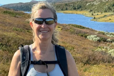 Frøydis Adamson Frøydis Adamson standing outside on heath land, by a lake - wearing sunglasses, a short sleeve top and rucksack straps are visible. She has blond hair and is smiling.