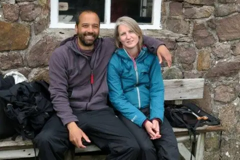 Tarig and Jenny sit on a bench together, smiling. Tarig's arm is around Jenny's shoulders. Tarig has a beard and is wearing a purple zip-top. Jenny has straight blond hair and is wearing a blue zip-top.