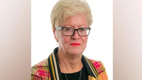 PA Helen Pitcher has short blonde hair and a red green and yellow tartan jacket on with a black collar with yellow stripes on it. She is wearing glasses. 