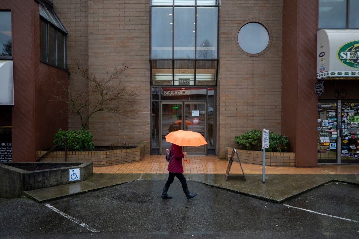 A person wearing a red coat and carrying an orange umbrella walks in the rain in front of a brick building.