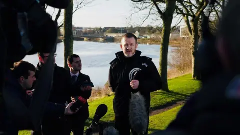 Police officer - Supt Davie Howieson - in uniform next to a river, surrounded by reporters, some holding recording devices.