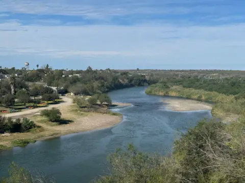 Bernd Debusmann/ BBC News Trees and some small buildings are on the left bank of a shallow river, with wild brush on the right