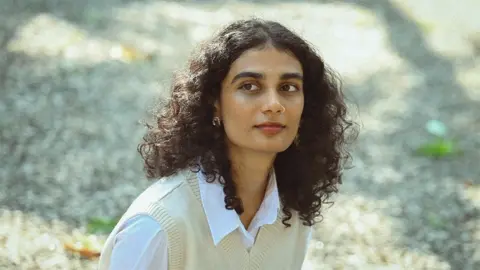 Gopan RS Anumita, wearing a cream coloured sweater vest over a white shirt, looking to the side while sitting outside.