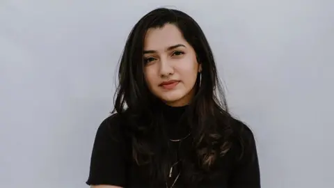 Kruthika Pillai Frizzell D'Souza, wearing a black top and shiny necklace. The background is plain light grey.