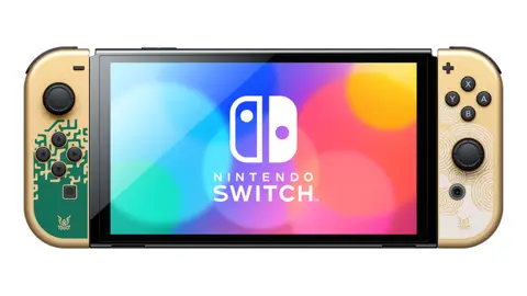 Nintendo A Nintendo Switch console against a white background. The screen displays the words "Nintendo Switch" and two icons representing the console's detachable controllers against a background of colorful circles creating a rainbow effect. The console's controllers are gold and printed with green and white patterns to represent the Legend of Zelda game series.