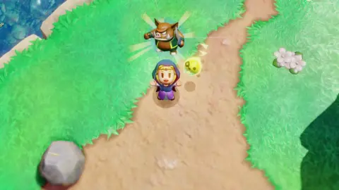 Nintendo Screenshot shows a top-down view of Princess Zelda wearing a navy blue cloak and standing on a dirt path surrounded by grass at the edge of a river. She's flanked by Tri - a floating, yellow, ball-shaped companion. She's looking at the sky, arms spread in a celebratory pose, and an icon of a humanoid pig enemy is floating above her head to show that it's just been captured.