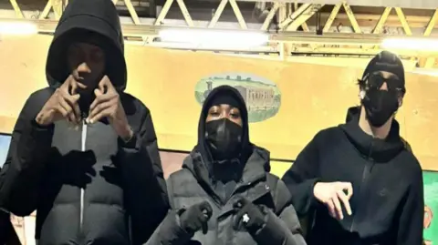 Met Police A photograph of Camron Osei, Shiloh Hinds and Bradley Harris who were all dressed in dark clothing, with masks and hoods. The trio are posing next to a graffitied sign next to Harrow and Wealdstone Tube station.