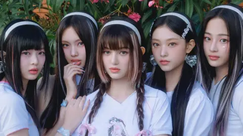 Siyoung Song / ADOR Promotional photo of the band NewJeans, with the five members dressed in white t-shirts, with white alice bands in their hair, against a floral backdrop.