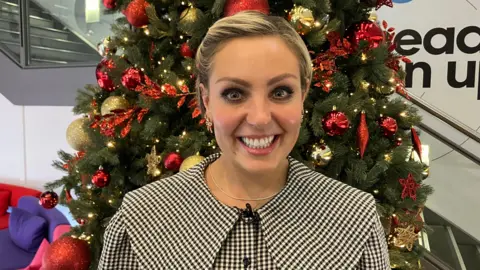 BBC Amy Dowden smiling looking into the camera. She is wearing chequered dress and a gold necklace is also visible. Behind her, a Christmas tree is decorated with baubles and festive lights.