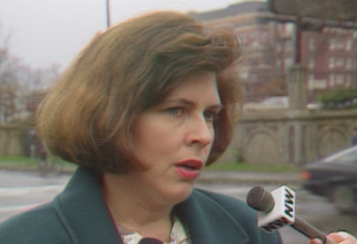 A woman with a brown bob haircut and pink lipstick speaks into several microphones. 