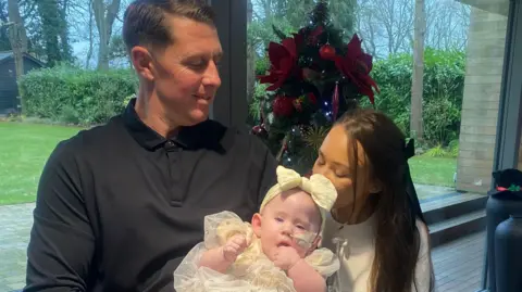 Supplied A man and a woman holding a baby girl. He is wearing a black top and she is wearing a white top. The baby is wearing a cream dress and matching bow. A decorated Christmas tree is in the background.