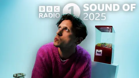 Ben Henratty Barry Can't Swim, dressed in a mauve jumper, looks upwards with pursed lips, in a light blue room dressed with a gumball machine and an old-school telephone.