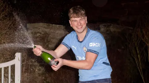 National Lottery James Clarkson in a garden shaking a bottle of Champagne which is spraying out of the bottle