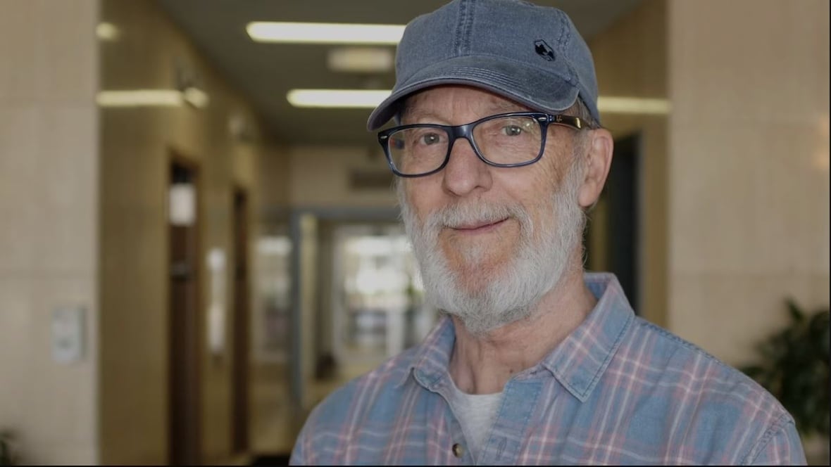 A man with a white beard, plaid shirt and blue hat, smiles at the camera with a hallway behind him. 