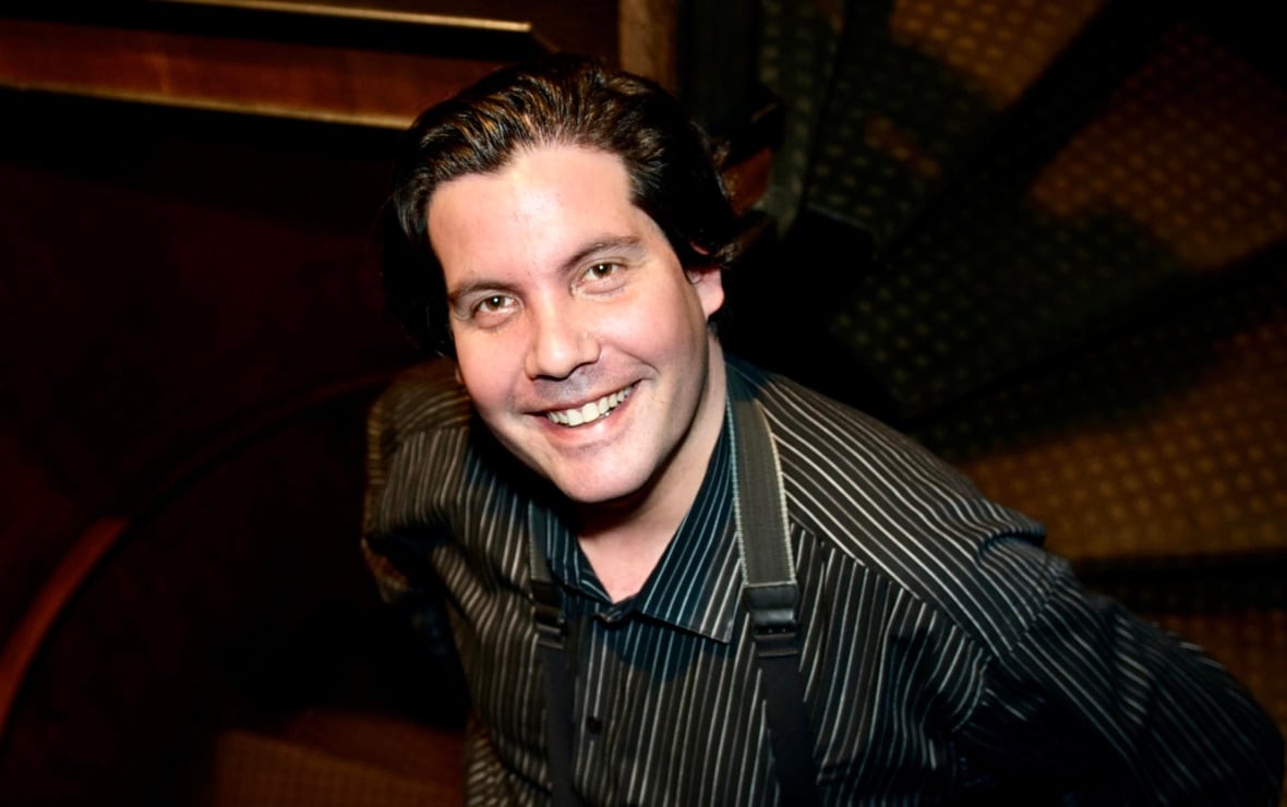 A man with black hair, a dress shirt and suspenders looks up at the camera with a smile. 