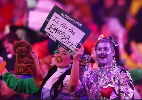 Reuters Fans in Wizard of Oz fancy dress - a woman dressed as Dorothy and a man dressed as the Tin Man - cheer at a darts match. The woman is holding a plushy dog toy and a sign saying "is this the Emerald City?" The man is holding a drink.