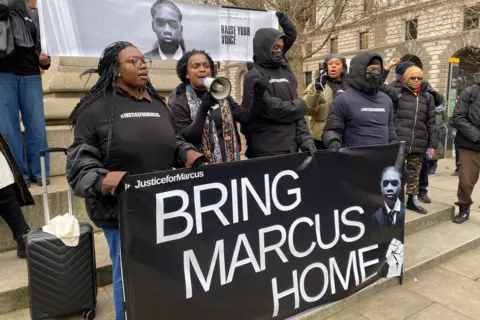 BBC About 10 campaigners holding a large black banner, with white lettering saying BRING MARCUS HOME. One person has a megaphone and they are wearing tops with 'justiceforMarcus' on.