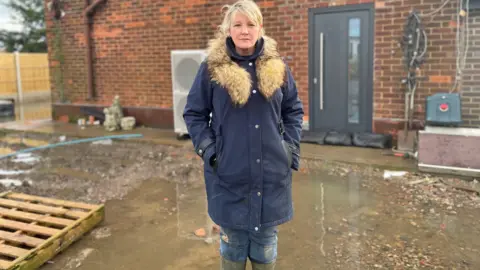 BBC/Simon Thake A woman with blonde hair stands in green wellies in flooded water.