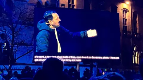 BBC Stephen Frayne shown on a big screen holding up a card