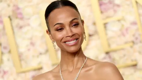 Getty Images Zoe Saldaña attends the 82nd Annual Golden Globe Awards at The Beverly Hilton on January 05, 2025 in Beverly Hills, California. She is smiling and is wearing a diamond necklace and matching drop earrings. 