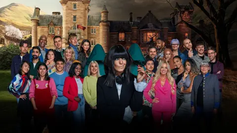 BBC/Studio Lambert Claudia Winkleman stands in front of 25 contestants against the backdrop of a grand Scottish castle/mansion in a publicity shot for The Traitors. Claudia throws a quizzical look from underneath her trademark fringe. She wears a black jacket over a white blouse, with wide cuffs that poke out from the sleeve ends.