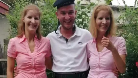 Huszti family Henrietta and Eliza Huszti standing either side of their brother. Both sisters have brown, shoulder-length hair and are wearing pink shirts. Their brother is in the middle and is wearing a white polo shirt and dark hat. 