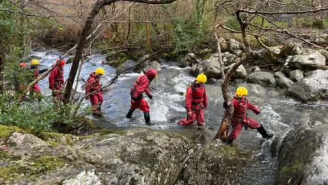 BBC A group of children walking through a rocky river gorge, wearing bright red safety suits, life jackets and yellow protective helmets.