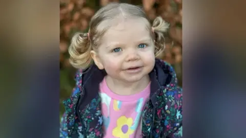 Gemma Whatling A young girl with blue eyes and blond hair tied up in a pig tail. She is wearing a colourful jacket and a pick jumper with flowers on