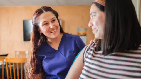 Getty Images Two women smiling at each other. One is a medical practitioner and is wearing blue scrubs, the other is a patient who seems to be being examined.