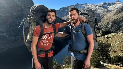 Family handout Aziz Ziriat, left, and Samuel Harris in shorts, t-shirt wirth large backpacks in front of a lake surrounded by mountains