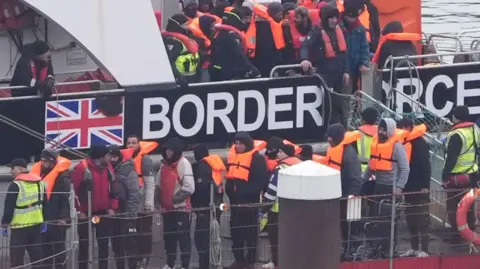 PA Refugees disembark from a Border Force boat. They are wearing orange life vests and are accompanied by officials.