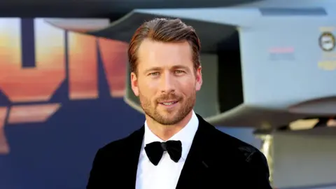 PA Media Glen Powell, dressed in a suit with bow tie, on the red carpet for a film premiere. He has dark brown hair and is unshaven with stubble. 