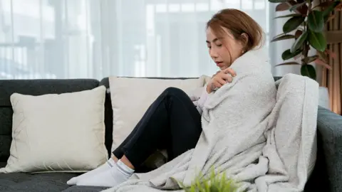 Getty Images A woman sits on the sofa with a blanket wrapped around her body to keep warm.