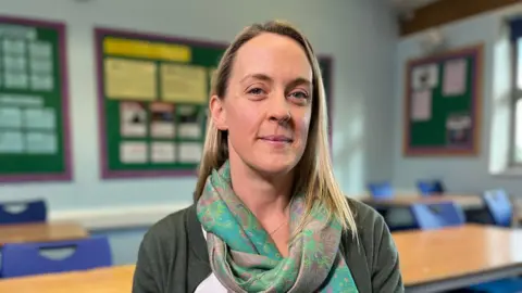 BBC A head and shoulders photo of a woman with straight blonde hair looking at the camera. She is wearing a green paisley scarf, olive cardigan and white blouse. She is sitting in a school classroom with wooden tables and blue chairs behind her, as well as display boards against the back wall.