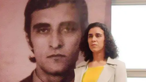 Tessa Moura Lacerda Tessa, a Brazilian woman wearing a yellow top, stands in front of a picture of her father