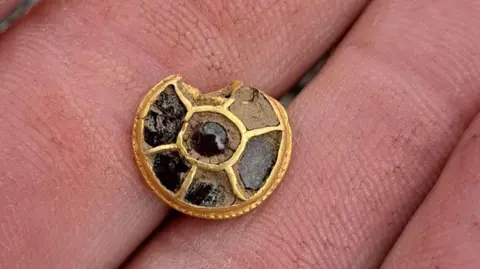 West Yorkshire Archaeology Advisory Service A small Anglo-Saxon era gold and red stone button or stud, slightly damaged on its top edge. It is round and has six compartments on its face, which were inlaid with red stones - some still existing. It is resting on the fingers of the detectorist who found it.